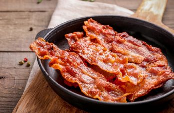Hot fried crispy bacon slices in skillet on wooden background.